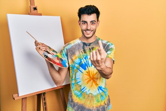 Young hispanic man standing drawing with palette by painter easel stand beckoning come here gesture with hand inviting welcoming happy and smiling