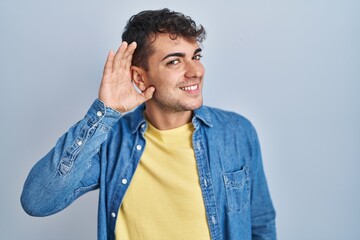 Young hispanic man standing over blue background smiling with hand over ear listening an hearing to rumor or gossip. deafness concept.