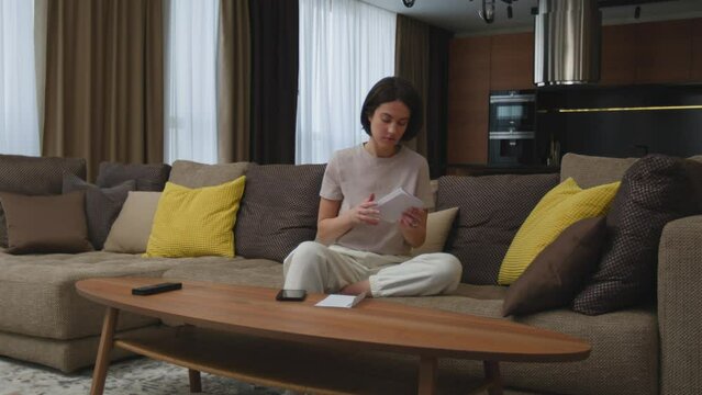 Upset female tenant homeowner looking through apartment bills sitting in living room at home, check financial taxes fees