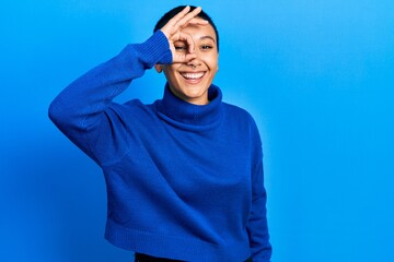 Beautiful hispanic woman with short hair wearing turtleneck sweater doing ok gesture with hand smiling, eye looking through fingers with happy face.