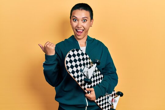 Beautiful hispanic woman with short hair holding skate pointing thumb up to the side smiling happy with open mouth