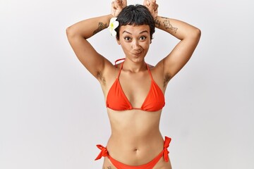 Young hispanic woman with short hair wearing bikini doing funny gesture with finger over head as...
