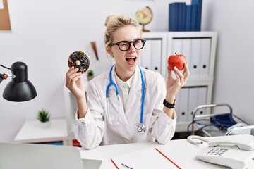 Young doctor woman holding red apple and donut at the clinic winking looking at the camera with...