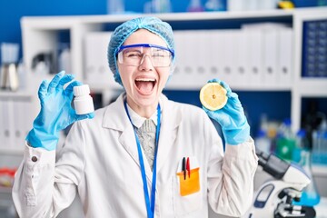 Beautiful woman working at scientist laboratory holding pills and lemon celebrating crazy and amazed for success with open eyes screaming excited.