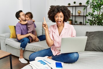 Mother of interracial family working using computer laptop at home pointing finger up with successful idea. exited and happy. number one.