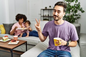 Hispanic father of interracial family drinking a cup coffee smiling looking to the camera showing fingers doing victory sign. number two.