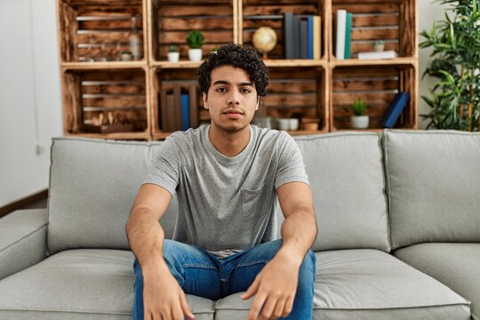 Young hispanic man wearing casual clothes sitting on the sofa at home with serious expression on face. simple and natural looking at the camera.
