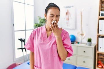 Young hispanic woman working at pain recovery clinic bored yawning tired covering mouth with hand. restless and sleepiness.