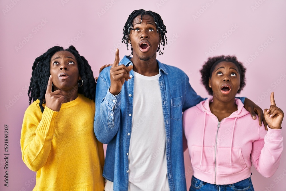 Canvas Prints Group of three young black people standing together over pink background amazed and surprised looking up and pointing with fingers and raised arms.