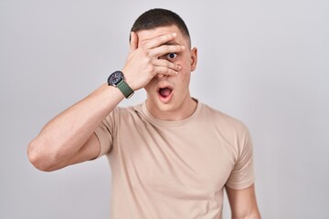 Fototapeta premium Young man standing over isolated background peeking in shock covering face and eyes with hand, looking through fingers with embarrassed expression.