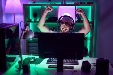 Young hispanic man streamer using computer and headphones with winner expression at gamin room