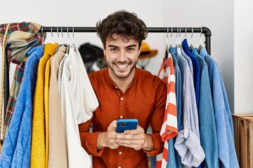 Young hispanic shopkeeper man smiling happy appearing through clothes using smartphone at clothing store.