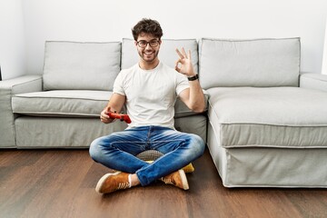 Hispanic man playing video game holding controller by the sofa doing ok sign with fingers, smiling friendly gesturing excellent symbol © Krakenimages.com