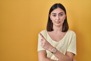 Obraz premium Hispanic girl wearing casual t shirt over yellow background pointing with hand finger to the side showing advertisement, serious and calm face
