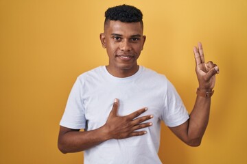 Young hispanic man standing over yellow background smiling swearing with hand on chest and fingers...
