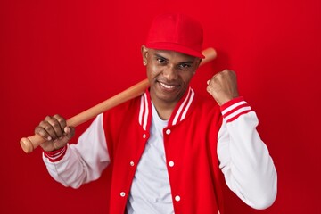 Young hispanic man playing baseball holding bat very happy and excited doing winner gesture with arms raised, smiling and screaming for success. celebration concept.