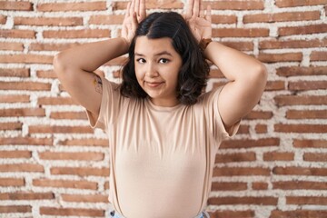 Young hispanic woman standing over bricks wall doing bunny ears gesture with hands palms looking cynical and skeptical. easter rabbit concept.