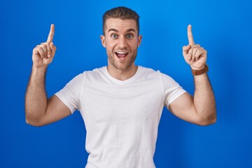 Fototapeta premium Young caucasian man standing over blue background smiling amazed and surprised and pointing up with fingers and raised arms.
