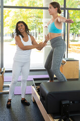 Personal female trainer controlling movements of young woman doing pilates on reformer in fitness studio