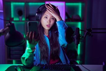 Young asian woman playing video games with smartphone surprised with hand on head for mistake, remember error. forgot, bad memory concept.