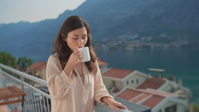 Young inspired asian woman standing terrace, enjoying drinking hot coffee, tea, watching views of sunrise landscape and scenery. People at home. Peaceful morning old European city in mountains by sea.