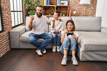 Family of three playing video game sitting on the sofa pointing thumb up to the side smiling happy with open mouth