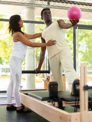 Experienced Latin female instructor helping African-American man during training with pilates bender ball to strengthen muscles in fitness studio