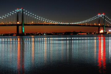 The lights string the Throgs Neck Bridge in Queens, New York City, and are reflected in the calm water below - obrazy, fototapety, plakaty