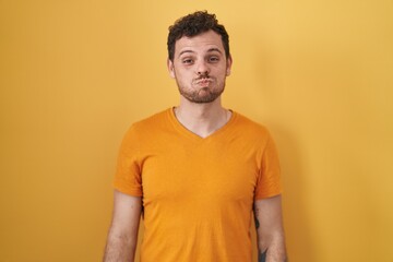 Young hispanic man standing over yellow background puffing cheeks with funny face. mouth inflated with air, crazy expression.