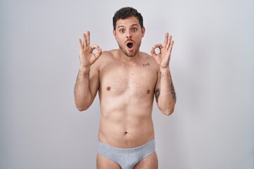 Fototapeta premium Young hispanic man standing shirtless wearing underware looking surprised and shocked doing ok approval symbol with fingers. crazy expression