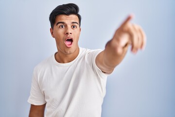 Hispanic man standing over blue background pointing with finger surprised ahead, open mouth amazed expression, something on the front