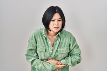 Young asian woman standing over white background with hand on stomach because indigestion, painful illness feeling unwell. ache concept.