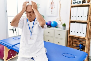 Senior physiotherapy man working at pain recovery clinic suffering from headache desperate and stressed because pain and migraine. hands on head.