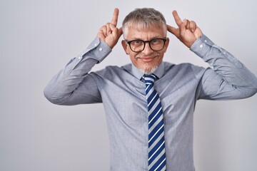 Hispanic business man with grey hair wearing glasses posing funny and crazy with fingers on head as bunny ears, smiling cheerful