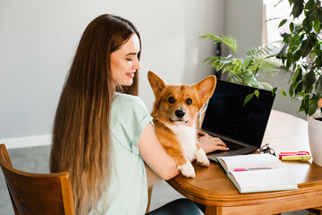 Distance education. Girl student with laptop study online with her lovely Corgi dog at home. Young woman hug Welsh Corgi Pembroke and making notes in notebook. Lifestyle with domestic pet.