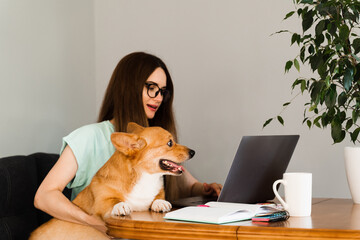 Distance education. Girl student in glasses with laptop study online with her lovely Corgi dog at home. Young woman hug Welsh Corgi Pembroke. Lifestyle with domestic pet