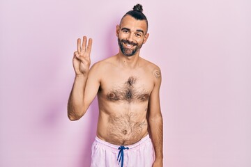 Fototapeta premium Young hispanic man shirtless wearing swimsuit showing and pointing up with fingers number three while smiling confident and happy.