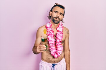 Young hispanic man wearing swimsuit and hawaiian lei drinking tropical cocktail looking sleepy and...