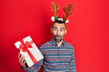 Young hispanic man wearing deer christmas hat holding gift scared and amazed with open mouth for...