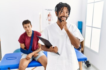 Obraz premium Young hispanic man working at pain recovery clinic with a man with broken arm with hand on chin thinking about question, pensive expression. smiling and thoughtful face. doubt concept.