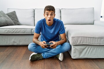 Young handsome hispanic man playing video game sitting by the sofa scared and amazed with open...