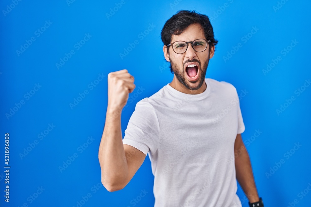 Poster Handsome latin man standing over blue background angry and mad raising fist frustrated and furious while shouting with anger. rage and aggressive concept.