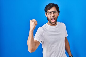 Handsome latin man standing over blue background angry and mad raising fist frustrated and furious while shouting with anger. rage and aggressive concept.