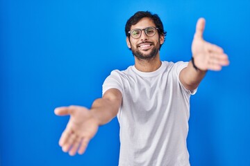 Naklejka premium Handsome latin man standing over blue background looking at the camera smiling with open arms for hug. cheerful expression embracing happiness.
