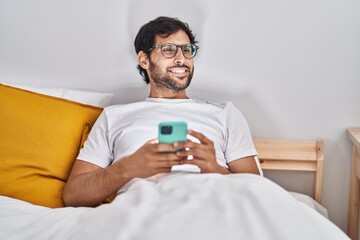 Young hispanic man using smartphone sitting on bed at bedroom