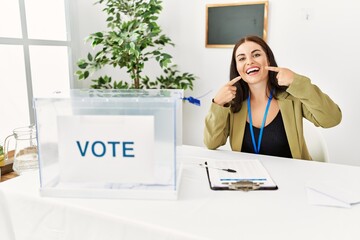 Young brunette woman sitting at election table with voting ballot smiling cheerful showing and...