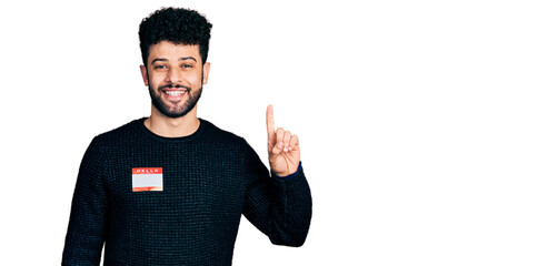 Young arab man with beard wearing hello my name is sticker identification smiling with an idea or question pointing finger with happy face, number one