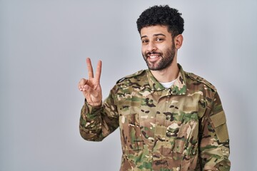 Arab man wearing camouflage army uniform smiling looking to the camera showing fingers doing victory sign. number two.