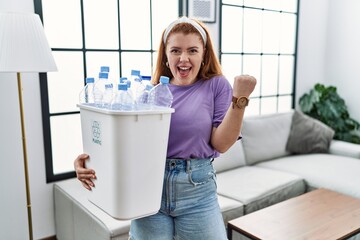 Young redhead woman holding recycling wastebasket with plastic bottles very happy and excited doing winner gesture with arms raised, smiling and screaming for success. celebration concept.