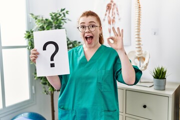 Young redhead physiotherapist woman holding question mark at the clinic doing ok sign with fingers,...